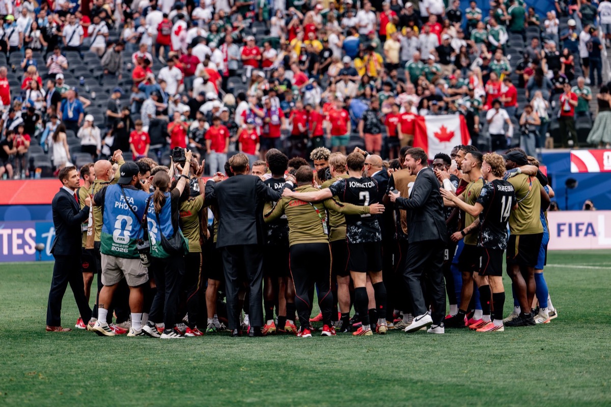 Football team in a huddle on the pitch after a match, fans cheering in the stadium behind them with a Canadian flag visible in the crowd