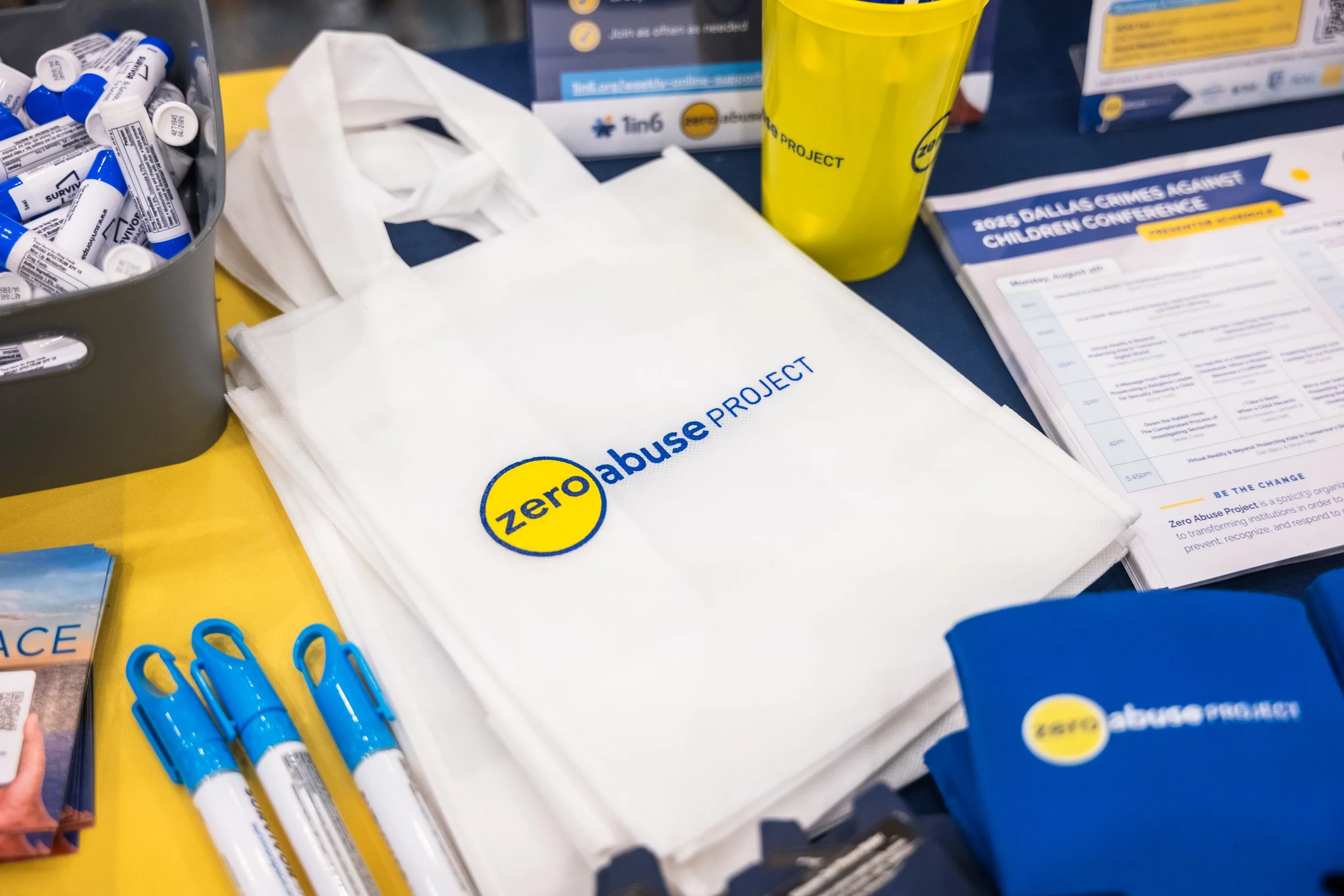 A table featuring a tote bag with the words 'Zero Abuse Project' and a yellow circle logo, blue and white pens, a yellow water cup, and informational pamphlets related to the Zero Abuse Project.
