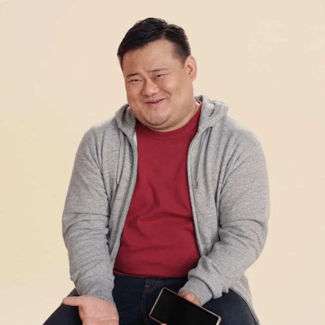 Individual sitting while holding a Pixel device, in a grey hoodie and red shirt, on a beige backdrop.