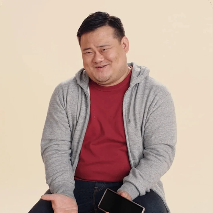 Individual sitting while holding a Pixel device, in a grey hoodie and red shirt, on a beige backdrop.